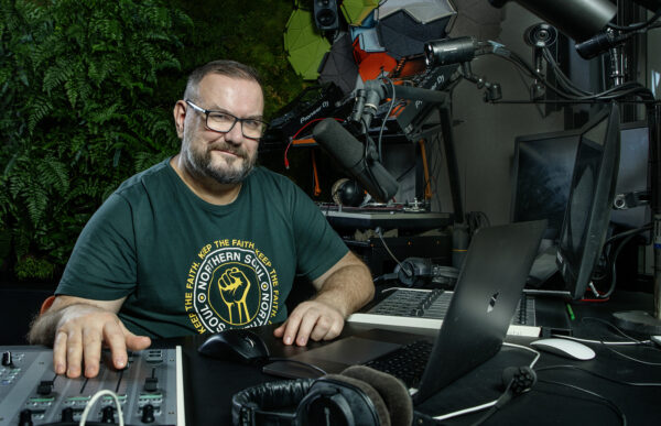 Njassa radiostudiossa t-paita päällä. Katsoo kameraan ja oikealla kädellä säätää äänilaitteita.