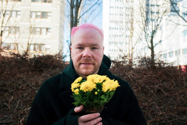 Haapa katsoo kameraan, pitelee käsissään keltaisia kukkia. Taustalla kerrostaloja.